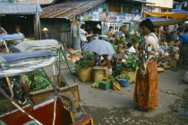 Market with sellers sitting around baskets of green vegetables with a market overseer looking on.