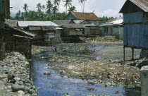 Wani. Housing next to street with river running through it contaminated with rubbish.  Near Palu