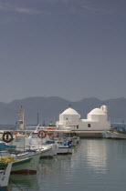 Fishing boats moored in Aegina Town next to a small white church with the mainland in the distance.