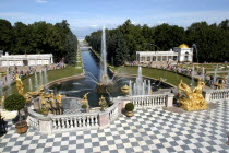 Peterhof Palace gardens. Stone balcony with chequered floor and golden statues with fountain beyond. Peterhof is also known as Petrodvorets.