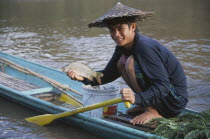 Fisherman  crouched on wooden canoe on the Mekong River holding up fish.
