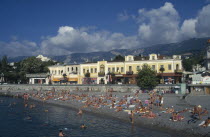 People sunbathing on narrow beach or swimming with promenade and waterside buildings behind.