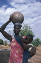 Woman breastfeeding child in sling at her side while carrying water pot on her head.