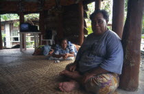 A Western Samoan tribal chief or Matai sitting on matting on floor inside house with television behind.