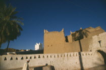 Man walking past whitewashed wall with mud brick town walls and white mosque minaret behindGadames