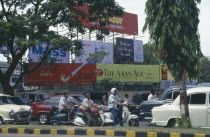 Street scene with cars and mopeds passing advertising hoardings.