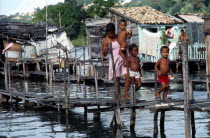 Young woman with three children crossing wooden bridge over water polluted with sewage in shanty town.  Houses raised on stilts above water behind.Brasil slum favela Brazil