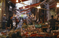 Vucciria Market.  Narrow street lined with fruit and vegetable stalls.  Passing pedestrians and moped riders.
