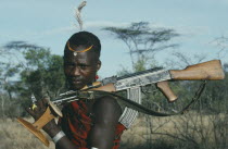 Portrait of Karamojong warrior with ostrich feather in hair holding gun and stool  always carried and used as both seat and head rest. Pastoral tribe of the Plains Nilotes group related to the Masi
