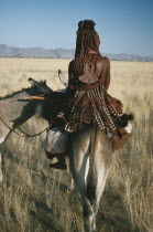 Himba tribeswoman on donkey seen from behind wearing typical leatherwork decorated with cowrie shells and copper.Semi nomadic pastoral people related to the Herero and speaking the same language