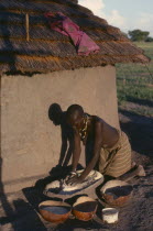 Dinka woman grinding dura outside hut casting shadow on wall.