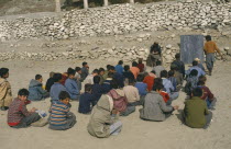 Outdoor classroom for boys only with teacher seated beside blackboard.