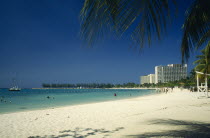 Sandy beach with people in shallow water near the shore and high rise apartments behind.  Palm fronds casting shadow in the foreground.