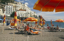 Amalfi.  Beach with sunbathers on loungers under yellow and orange striped beach umbrellas.  Town buildings on the hillside behind.