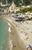Amalfi.  View along shingle beach with sunbathers and cafes towards town buildings on hillside.