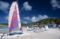 Sandy beach with people on deck chairs beside blue painted building  A-board advertising Tonys watersports.  Hobiecat with bright sail in front.
