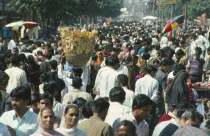 Crowded street scene with man in centre carrying basket of bread on his head.