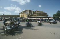 Trishaws and motorcyclists on road in front of the Central market buildings.
