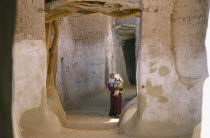 Woman carrying child along street between mud brick buildings.