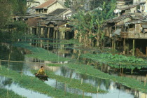 Wooden houses raised on stilts above waterway with man in small canoe in foreground.