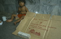 Homeless child sitting on ground next to carboard box with a polite begging note written in English on it