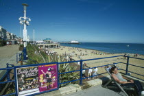 View from promenade towards beach and bandstand with a woman sat on deckchair next to blue railings in the forground.