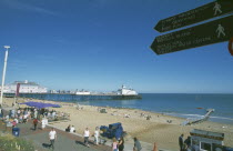 View across busy seafront towards shingle beach and pier with direction signs for tourist attractions in the forground.