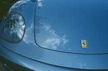 Detail of blue Ferrari emblem and front light with clouds reflected in paintwork.
