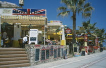 Promenade with English restaurant and bar signs. People sat at tables.Costa del Sol Andalusia