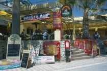 Promenade with English restaurant and bar signs. People sat at tables.Costa del Sol
