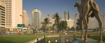 Camel statue in Al-Ittihad Square  people sat on grass. Palm trees and car park in distance.Travel  Tourism  Panorama  Dubayy Automobile  United Arab Emirates