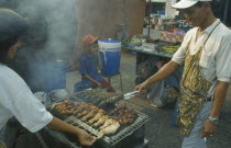 Man cooking fish and skewered meat over barbeque at roadside food stall with young boy sitting on stool behind.