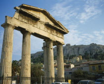 Plaka - Ancient Agora and Acropolis on top. Old ruined columns behind a fence with houses and a hill in the background.