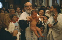 A Greek orthodox christening  the child being held up by the Priest with friends and family behind.