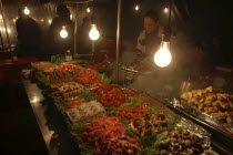 Namdaemun Market on a cold December night with a woman cooking behind trays of fresh seafood  pork  chicken and beef with plastic sheeting to keep out cold