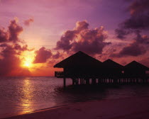 View of silhouetted beach huts on stilts over water against a dramatic pink setting sky