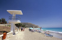 Cyprus, Pissouri Bay, sunbathers on narrow sand and pebble beach with people walking along promenade at side and life guards tower or look out point in the foreground.