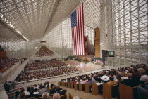 Crystal Cathedral Memorial Service. Congregation in pews under glass structure beneath flag of Stars And Stripes and a large TV