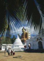 Site of giant seated Buddha constructed in 1970 with visiting family in courtyard in foreground.  Part framed by palm fronds.Vihara meaning image house