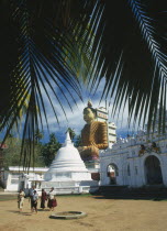 Site of giant seated Buddha constructed in 1970 with visiting family in courtyard in foreground.  Part framed by palm fronds.Vihara meaning image house