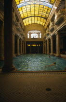 Gellert Baths.  Interior with bathers in swimming pool lined with carved pillars and with domed glass roof above.