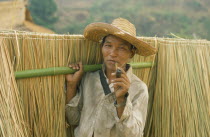 Akha man smoking bamboo pipe whilst carrying grasses collected for roofing