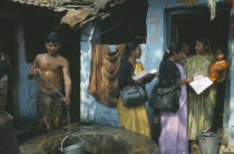 Women conducting a survey on reproductive health talk to a woman holding a child standing in the doorway of a house.  Man collecting water from well in the foreground.