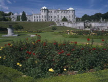 View over formal gardens towards exterior facade.  Flowerbed with red and yellow roses in the immediate foreground.