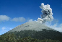 Smoke rising from the volcano as it erupts above the tree lined base with dust surrounding the summit
