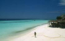 Nungwi.  Single figure walking along sandy beach beside aquamarine water.  Beach huts  tables and chairs on raised wooden platform on righthand side.