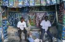 Tanzania, Zanzibar Island, arts and crafts, two young men in open fronted shop selling  paintings in the Tingatinga style depicting african animals in bold colours.