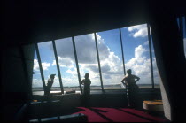 Miami Airport. Three figures looking out of window towards planes framed by interior walls in heavy shadow.
