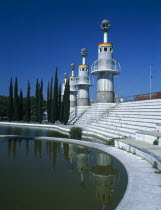 Parc Espanya Industrial.  Line of towers with balconies encircling their walls and spruce trees reflected in circular pond in the foreground.