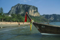 Long tail boat moored in shallow water at the edge of beach with tourists.  Forest covered cliffs behind.